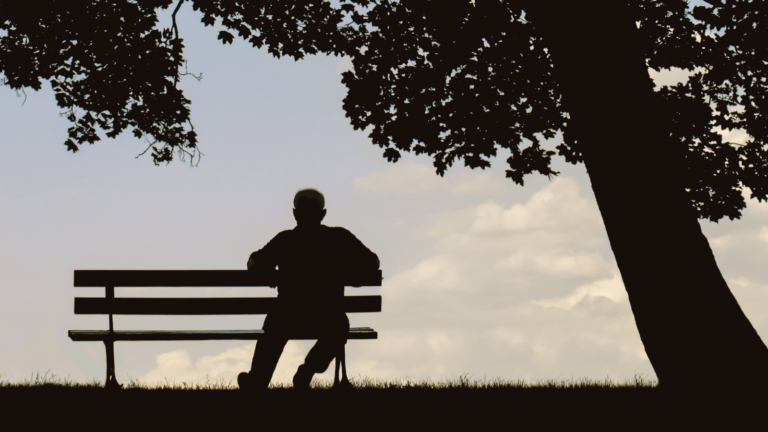 Mann sitzt bei Sonnenuntergang allein auf einer Parkbank unter einem Baum, symbolisiert Nachdenklichkeit und Trennungsschmerz.